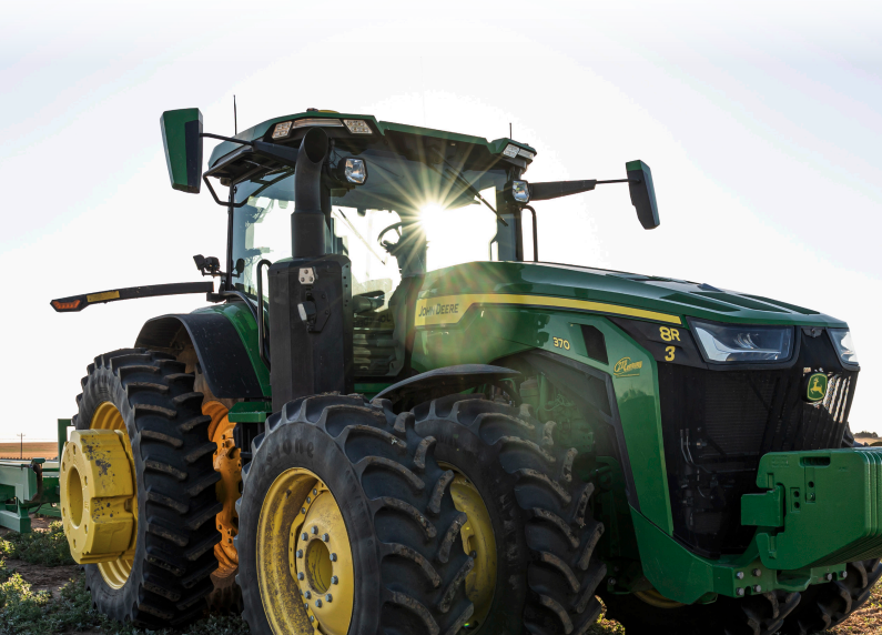 A green John Deere tractor in a field with the sun rising in the distance.